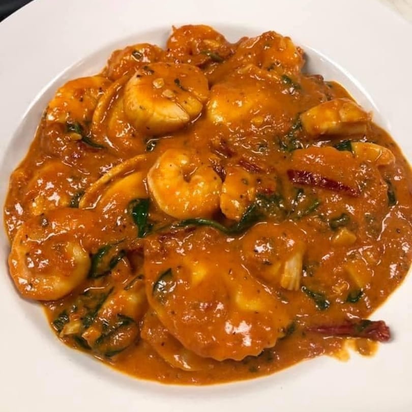 A white bowl filled with shrimp and ravioli in a rich orange-red sauce with green herbs.